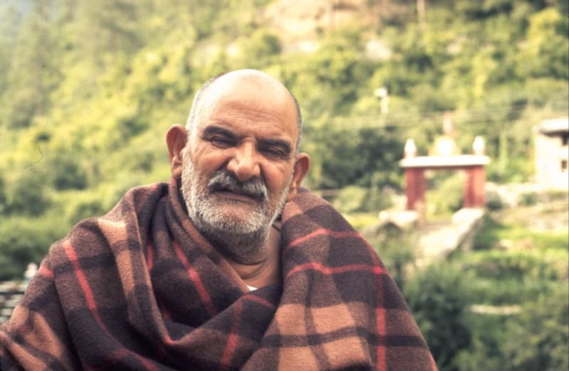 Neem Karoli Baba seated outdoors in a red plaid blanket on a green hillside