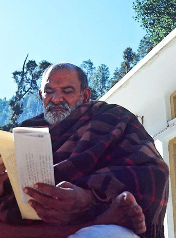Neem Karoli Baba wrapped in a blanket, reading a letter outside a hillside temple