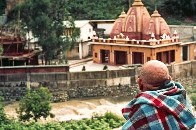 Neem Karoli Baba, wrapped in a blue and red blanket, looking across a river to a hillside temple