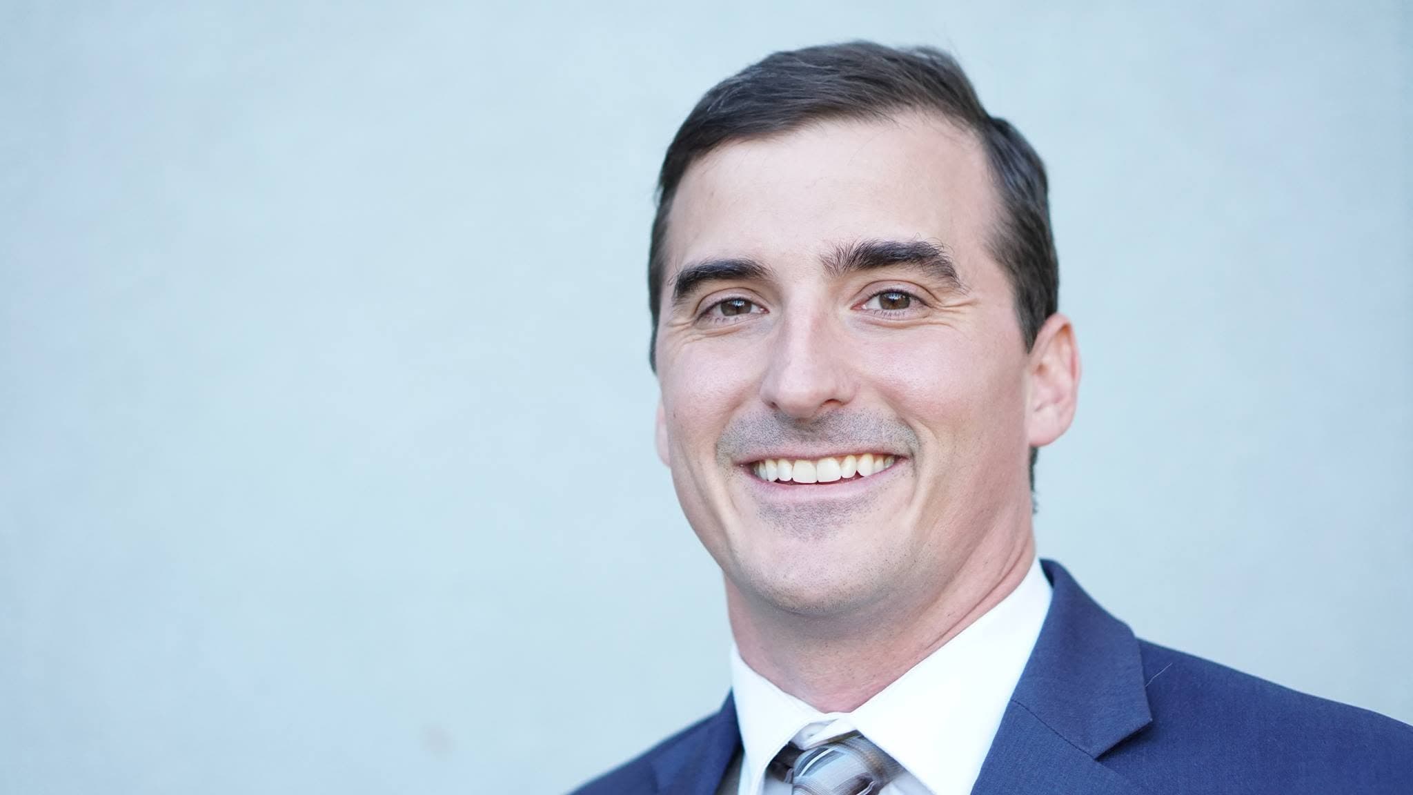 Tom smiling in a navy suit and tie against a pale blue background