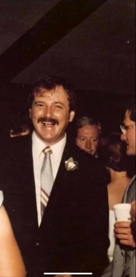 Thomas Whigham's father, a mustached man in a dark suit with a boutonniere, holds a cup at a party.