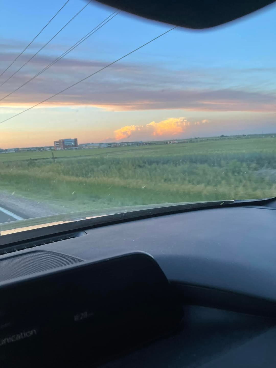 Sunset clouds over flat green fields seen through a car windshield, with power lines overhead.