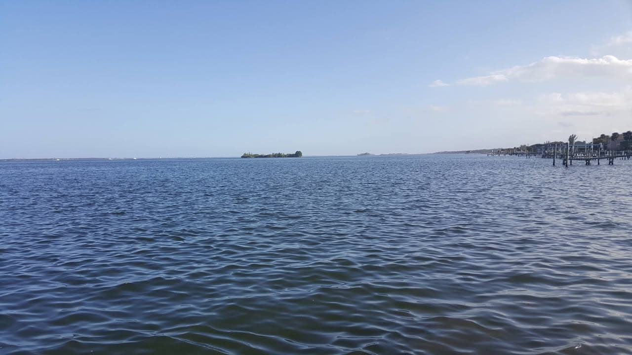 Calm blue bay water under a hazy sky, with a distant shoreline and dock pilings on the right.