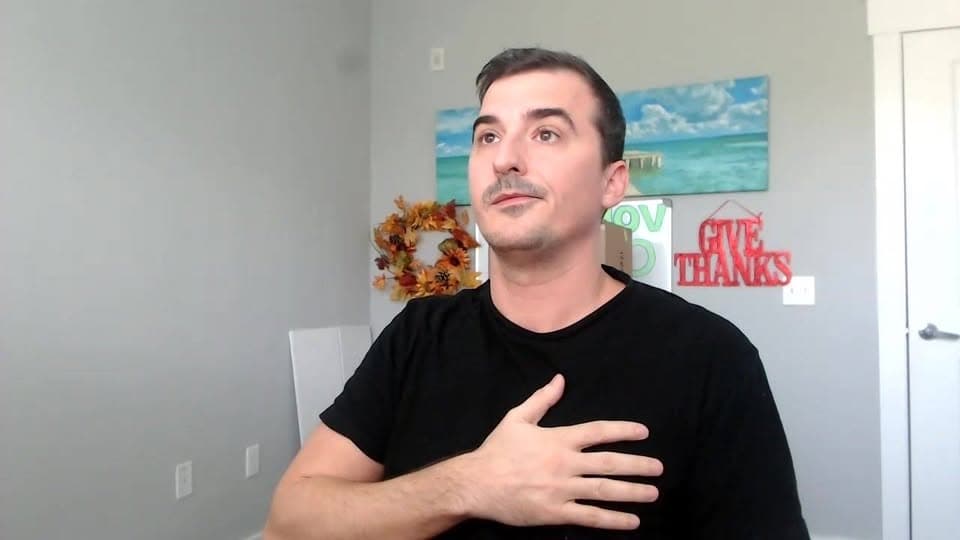 A man in a black T-shirt presses a hand to his chest while talking to the camera at home.
