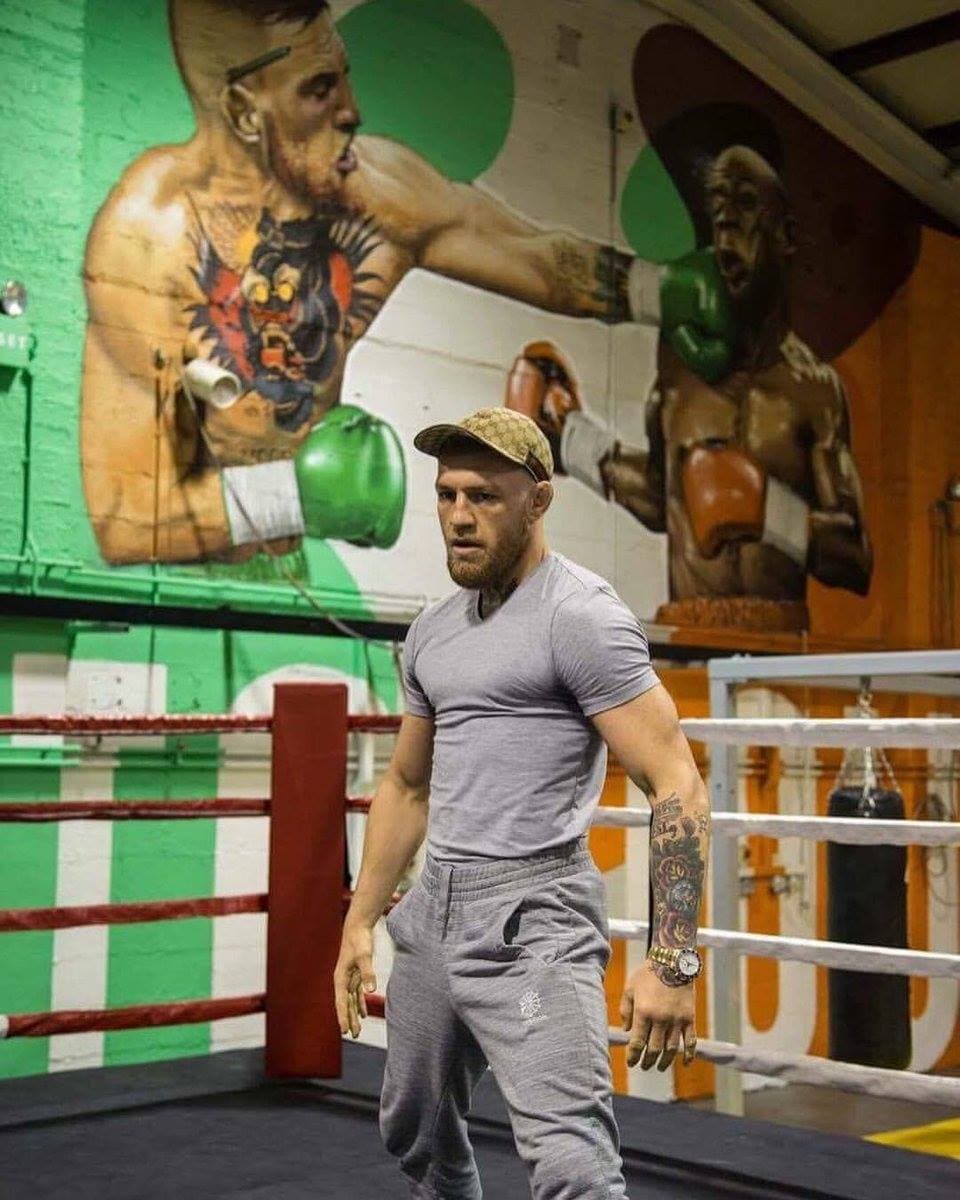 A heavily tattooed man in a grey T-shirt walks through a boxing gym past a mural of fighters.