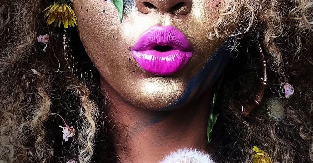 Close-up of a face dusted in gold glitter with bright magenta lips and flowers tucked into curly hair.