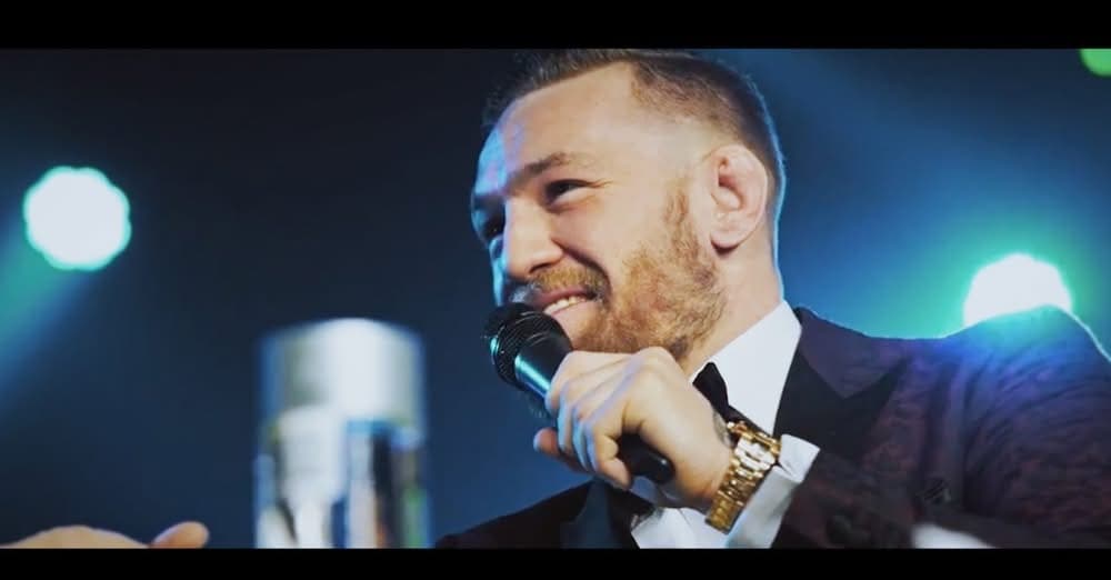A red-bearded man in a dark suit and gold watch speaks into a microphone under blue stage lights.