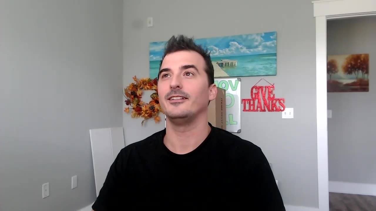 A man in a black T-shirt glances sideways in a living room with a beach painting and Give Thanks sign.