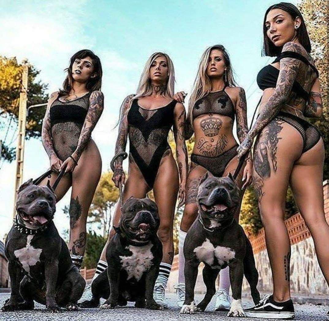 Four tattooed women in black outfits posing outdoors with three dogs.