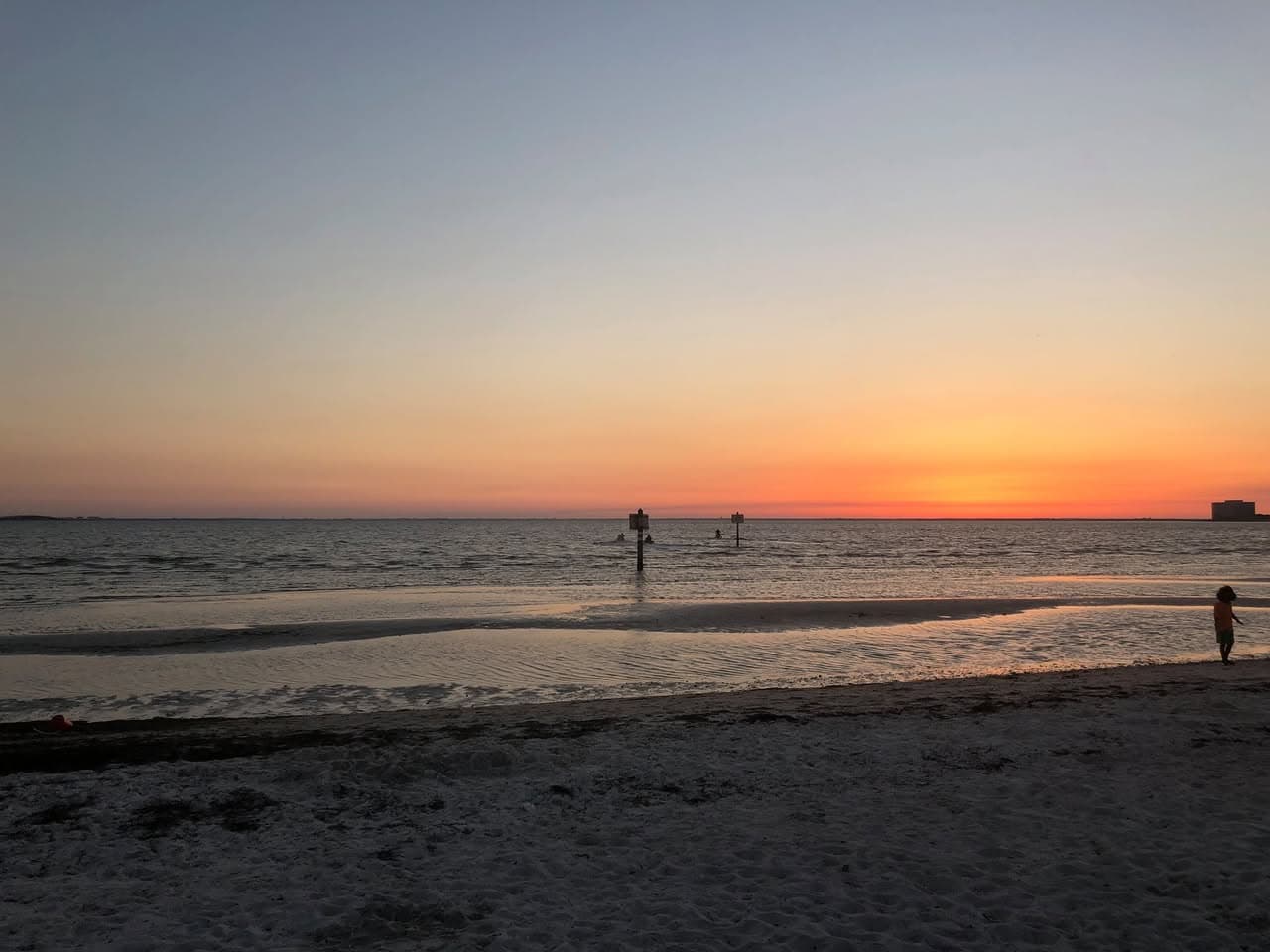 Orange sunset over a calm beach at low tide, with a lone figure standing at the water's edge.