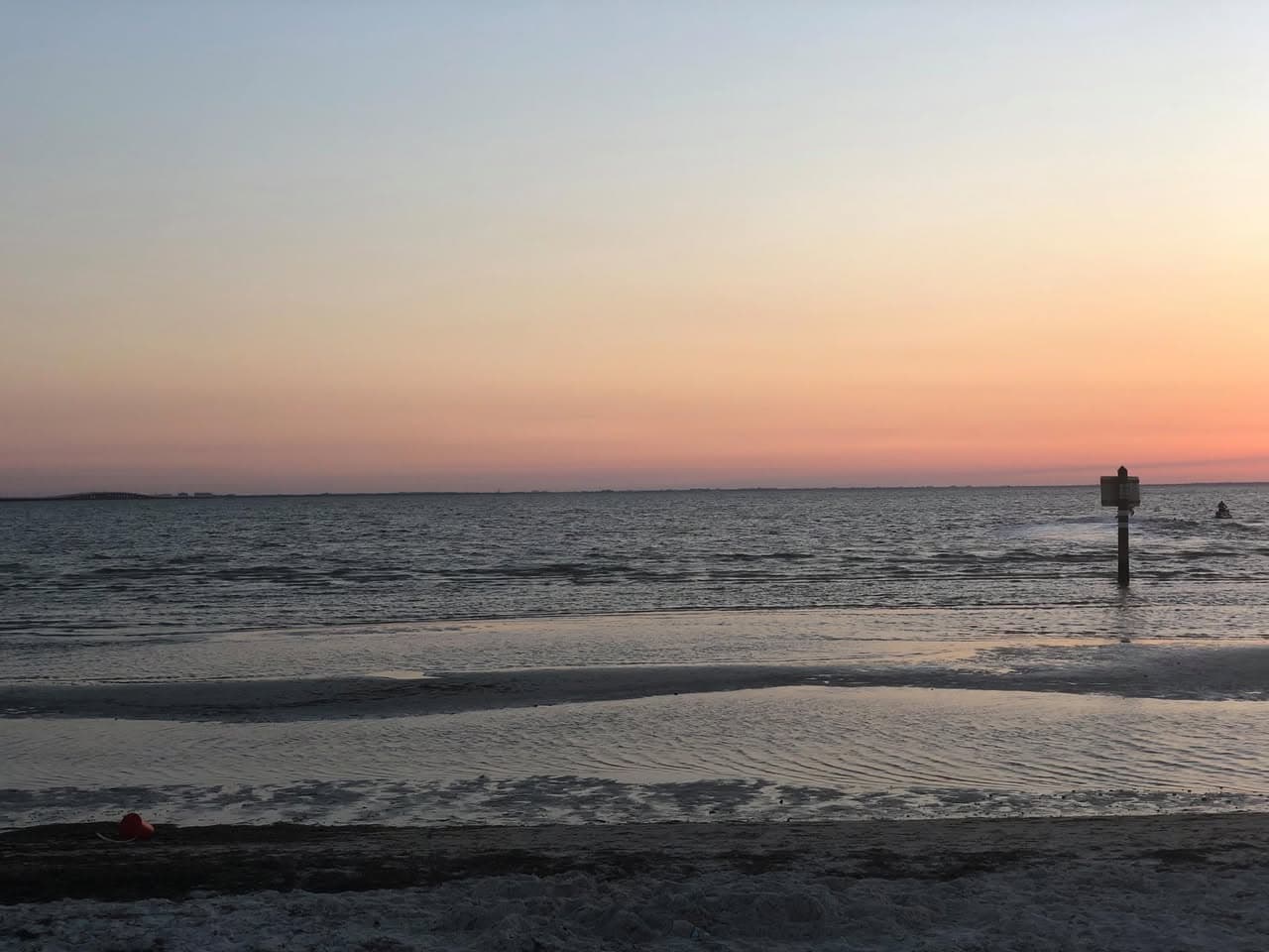 Pink and orange dusk over shallow water and wet sand, with a channel marker post offshore.