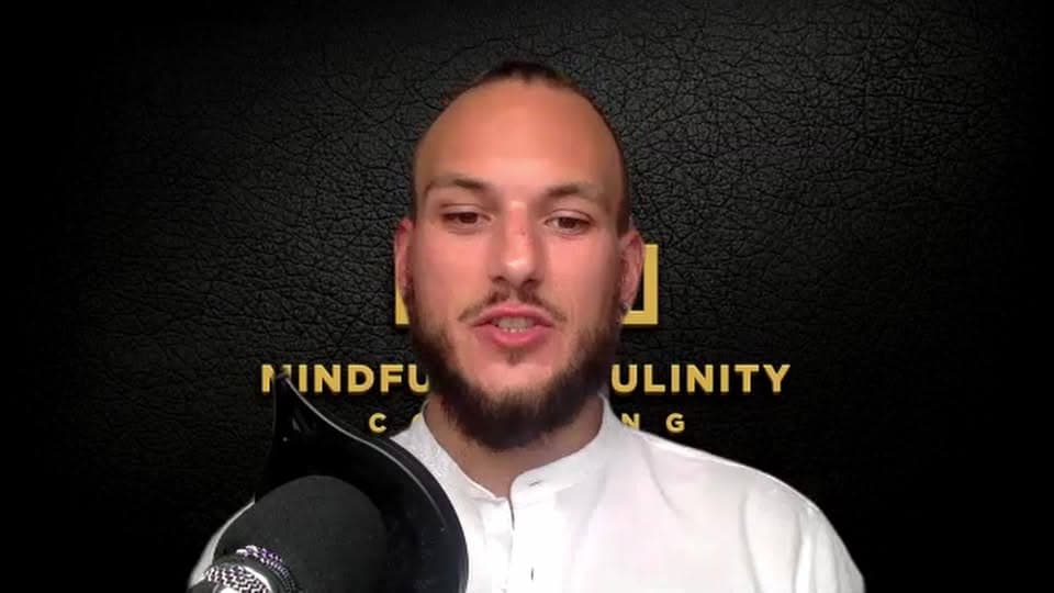 A bald, bearded man in a white shirt speaks into a microphone in front of a Mindful Masculinity Coaching logo.