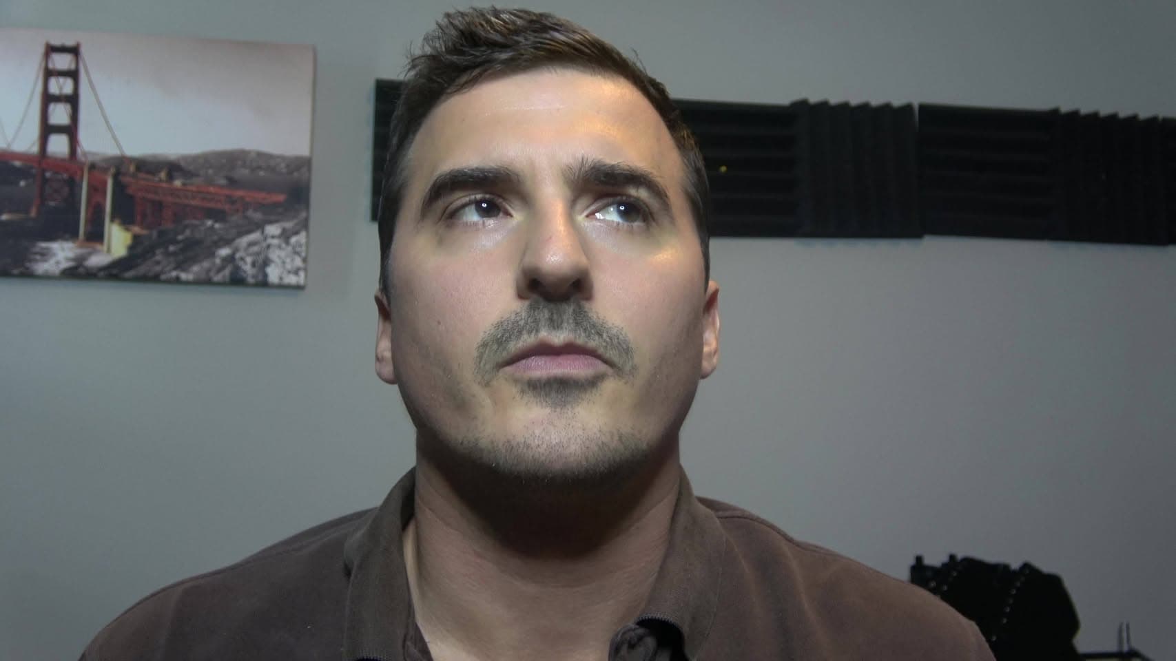 A man with a mustache looks upward in a room with a Golden Gate Bridge print and foam sound panels.