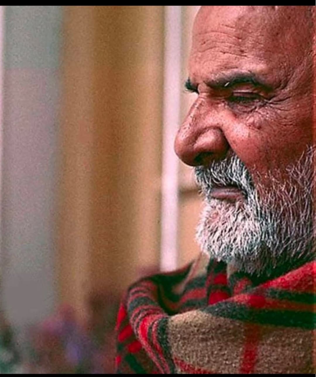 Close profile of Neem Karoli Baba, an elderly bearded man wrapped in a red and green plaid blanket.