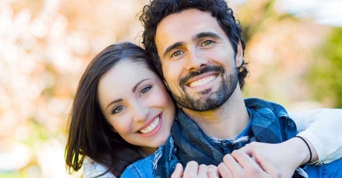 A smiling dark-haired woman and a bearded man pose cheek to cheek outdoors in autumn light.
