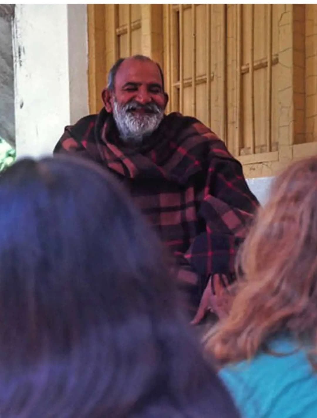 A smiling bearded man wrapped in a plaid blanket, seated before a group.