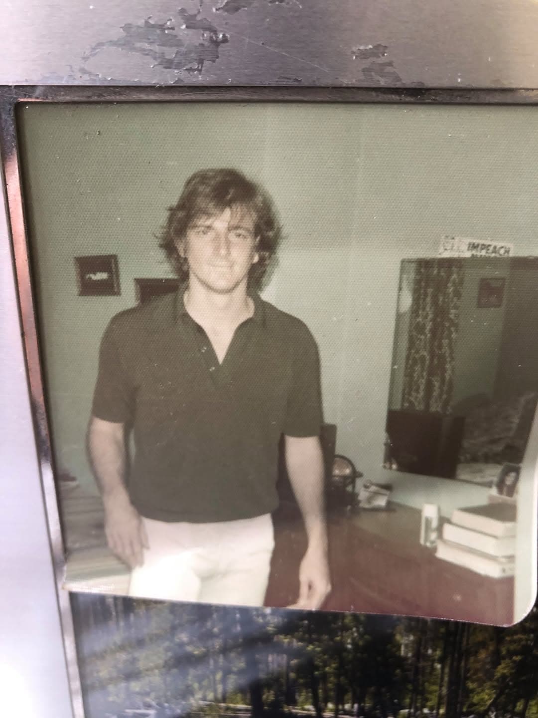 Faded vintage snapshot of a young man with shaggy hair in a dark polo shirt standing in a room.